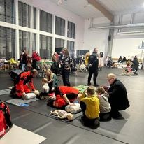 Grupa osób ćwiczy resuscytację krążeniowo-oddechową na manekinach w przestronnej sali z dużymi oknami. Instruktor w czerwonym kombinezonie demonstruje technikę, a uczestnicy powtarzają czynności.