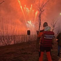 Ratownik medyczny w pomarańczowym kombinezonie z napisem 'RATOWNIK' na plecach, obserwujący pokaz sztucznych ogni w tle, z widocznym nagłośnieniem i drzewami bez liści.