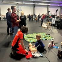 Instruktor w pomarańczowym uniformie demonstruje resuscytację krążeniowo-oddechową na manekinie niemowlęcia, podczas gdy mały chłopiec w niebieskiej bluzie naśladuje ruchy, w tle grupa obserwatorów...