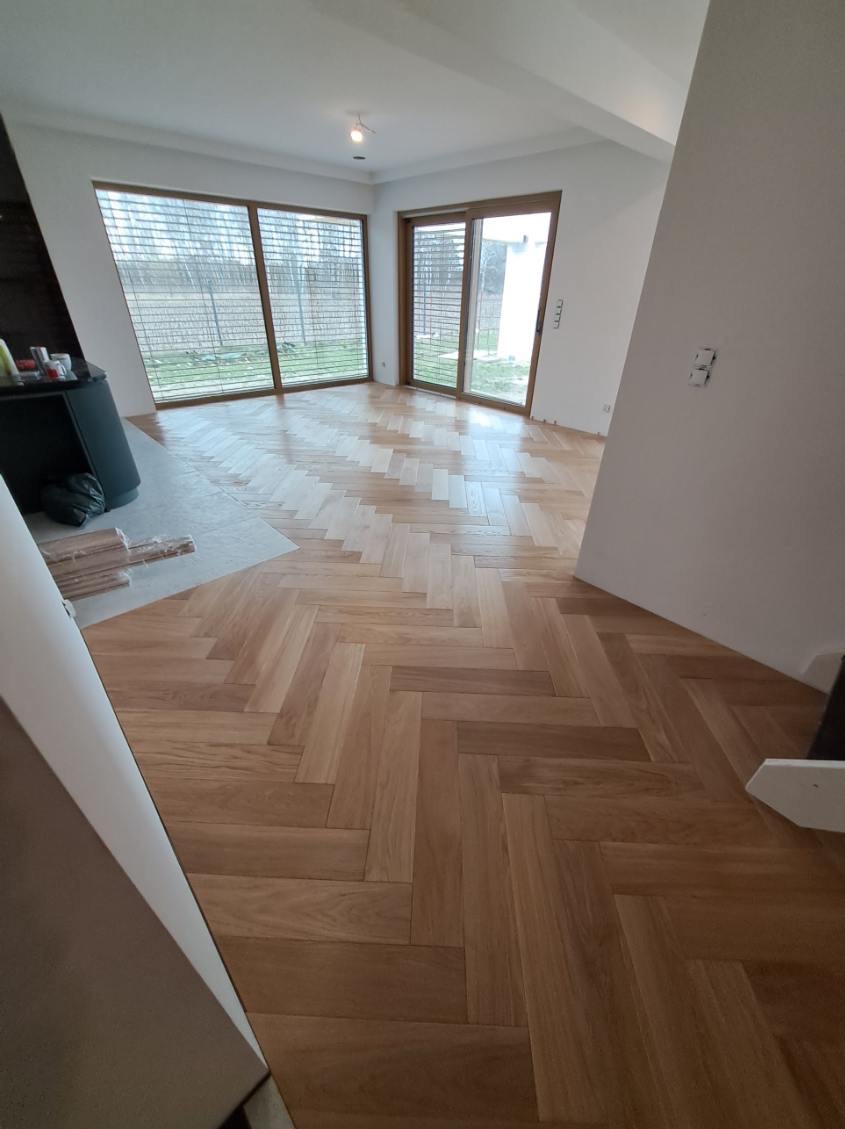 Jasna, drewniana podłoga ułożona w jodełkę w przestronnym, nowym wnętrzu z dużymi oknami i niedokończonymi detalami wykończeniowymi.
