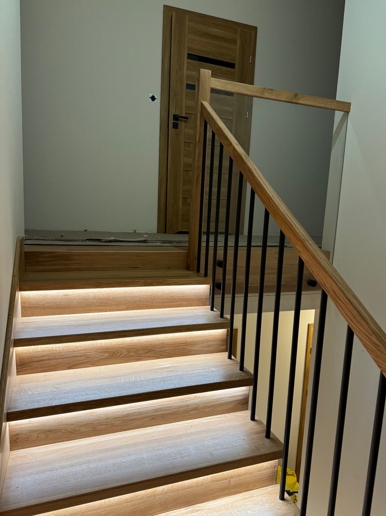 Oświetlone od spodu drewniane schody z czarnymi, metalowymi balustradami i szklanym panelem bocznym, prowadzące do drewnianych drzwi. Na górze schodów widoczne zabezpieczenia podłogi.