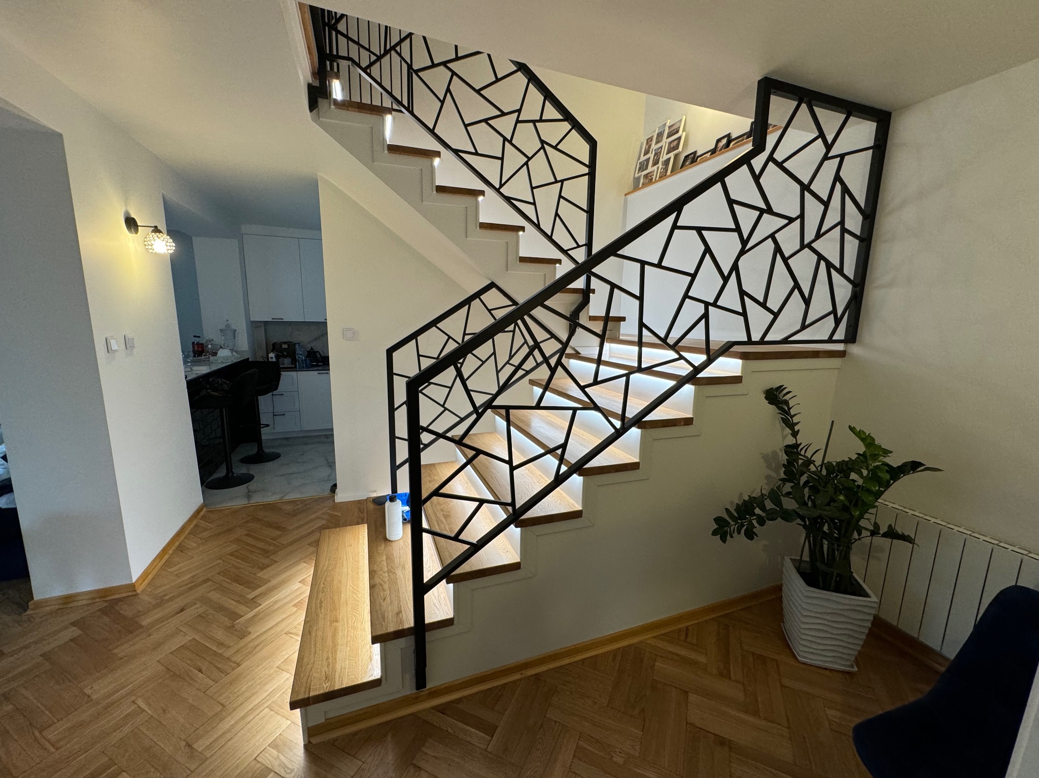 Nowoczesne schody z drewnianymi stopniami, podświetleniem LED i geometryczną, czarną balustradą, widok z dołu na tle parkietu w jodełkę.