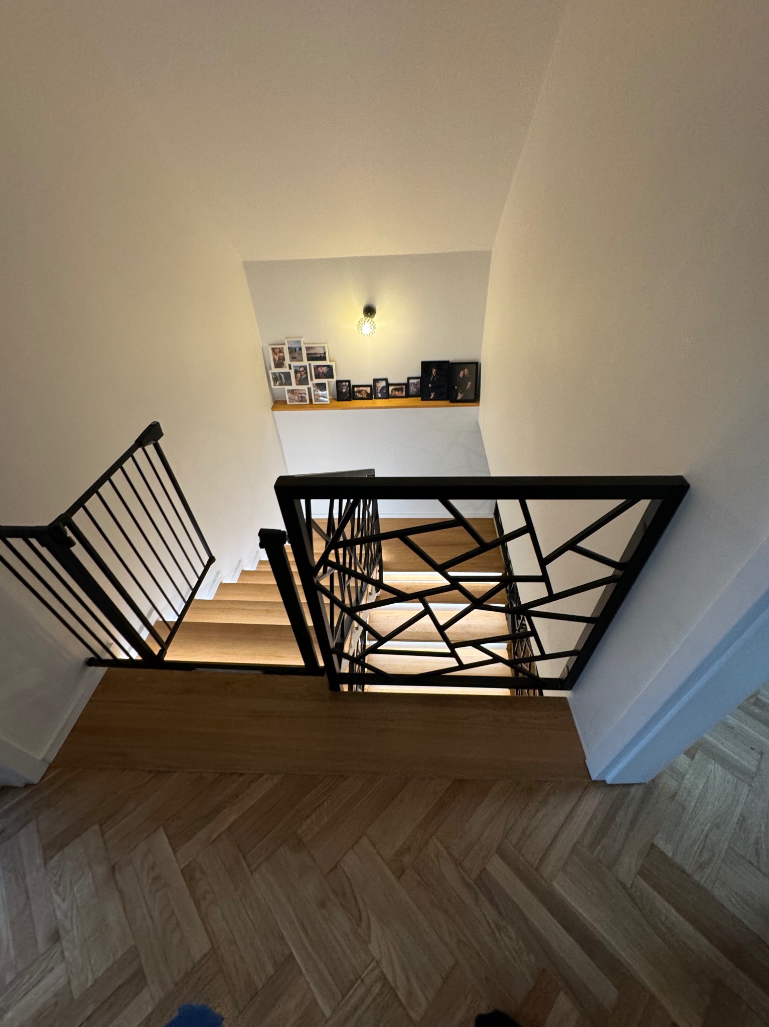 Widok z góry na dębowe schody z czarną balustradą o geometrycznym wzorze, parkiet ułożony w jodełkę i galerię zdjęć na ścianie w tle.