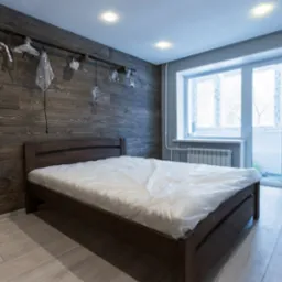 Sypialnia po remoncie: ściana z ciemnego drewna, ł&oacute;żko z przykryciem ochronnym, szare ściany, jasna podłoga i okno balkonowe.