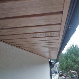 Łukasz Huzarski - Zamontowana podbitka dachowa w odcieniach brązu, widok z dołu. Widoczna ściana budynku z fakturą i fragment rynny. W tle zieleń drzew.