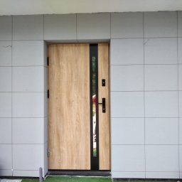 Łukasz Huzarski - Elewacja domu wykończona szarymi płytami imitującymi kamień, z drewnianymi drzwiami wejściowymi z czarnymi akcentami. Widoczne kwiaty w donicach i fragment trawnika.