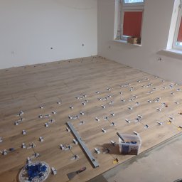 Łukasz Huzarski - Układanie paneli podłogowych w jasnym odcieniu drewna, z użyciem klipsów poziomujących. Widoczna poziomica i pojemnik z klipsami. Dwa okna z roletami w tle.