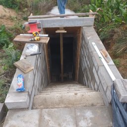 Łukasz Huzarski - Betonowe schody prowadzące w dół, z widocznymi narzędziami murarskimi i poziomnicą. W tle osoba w czerwonej bluzie i zielona roślinność.