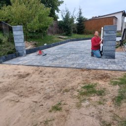 Łukasz Huzarski - Układanie kostki brukowej i montaż obrzeży w Stolno. Mężczyzna z poziomicą kontroluje pionowość słupka ogrodzeniowego z szarej kostki. Widoczne narzędzia i częściowo ułożony chodnik.