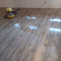 Łukasz Huzarski - Położona podłoga z paneli imitujących drewno, mokra po umyciu. W rogu żółty odkurzacz przemysłowy. Widoczne gniazdka elektryczne na ścianie.