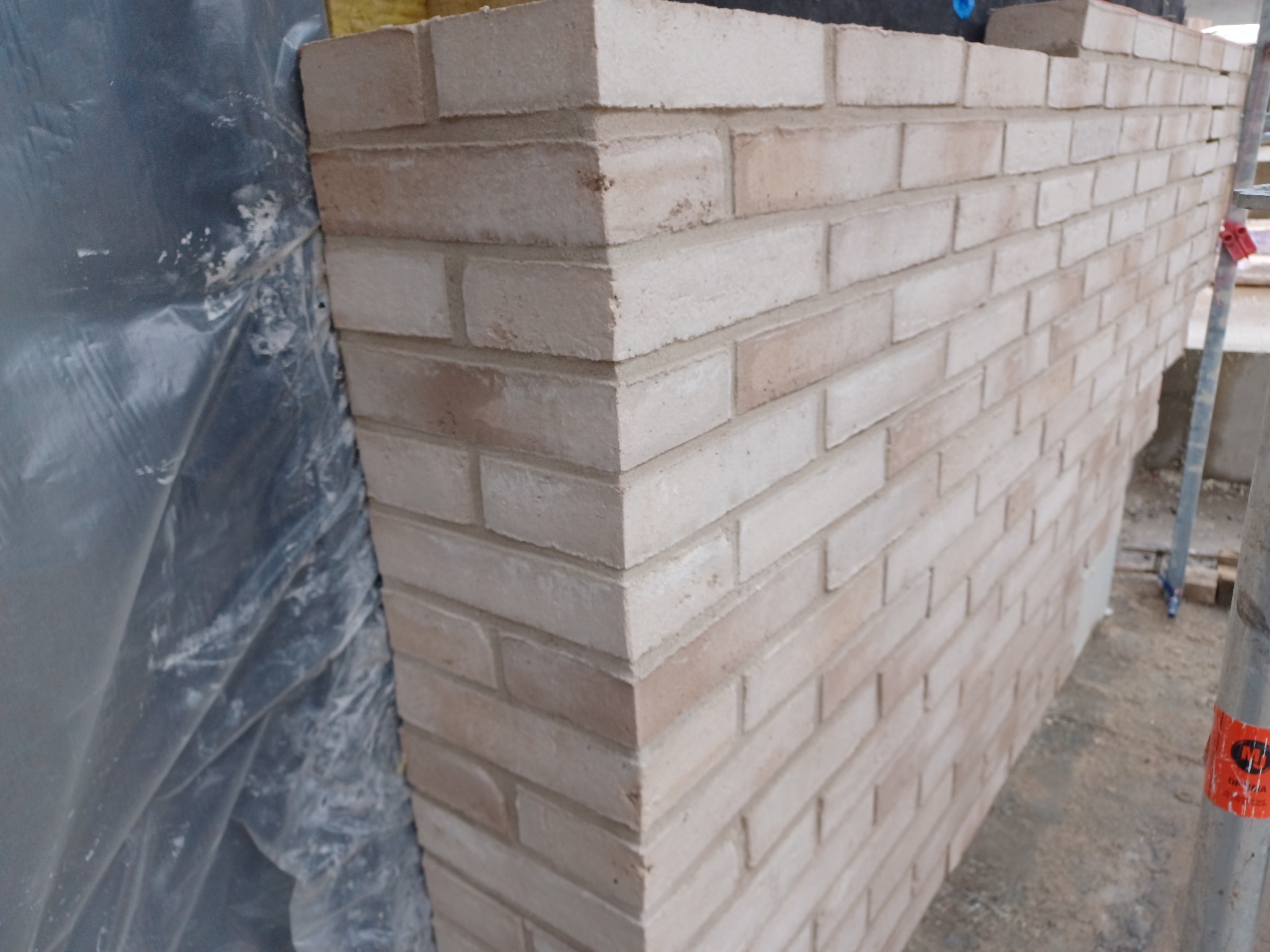 Fragment nowo wymurowanej ściany z jasnej cegły, widoczne rusztowanie i folia ochronna.