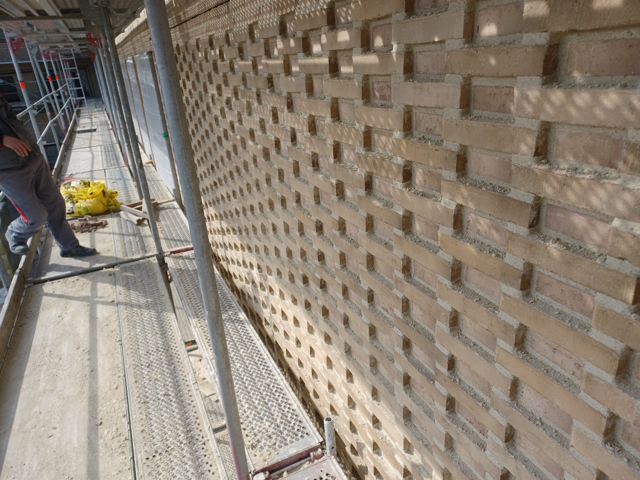 Ściana z cegieł ułożonych w dekoracyjny wzór, widoczna z rusztowania, z fragmentem sylwetki pracownika i żółtą torbą w tle.