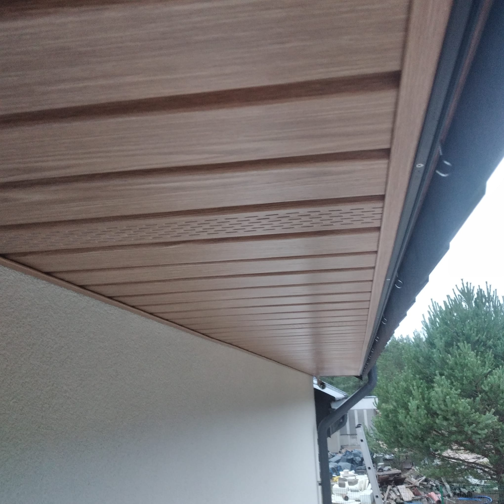Zamontowana podbitka dachowa w odcieniach brązu, widok z dołu. Widoczna ściana budynku z fakturą i fragment rynny. W tle zieleń drzew.
