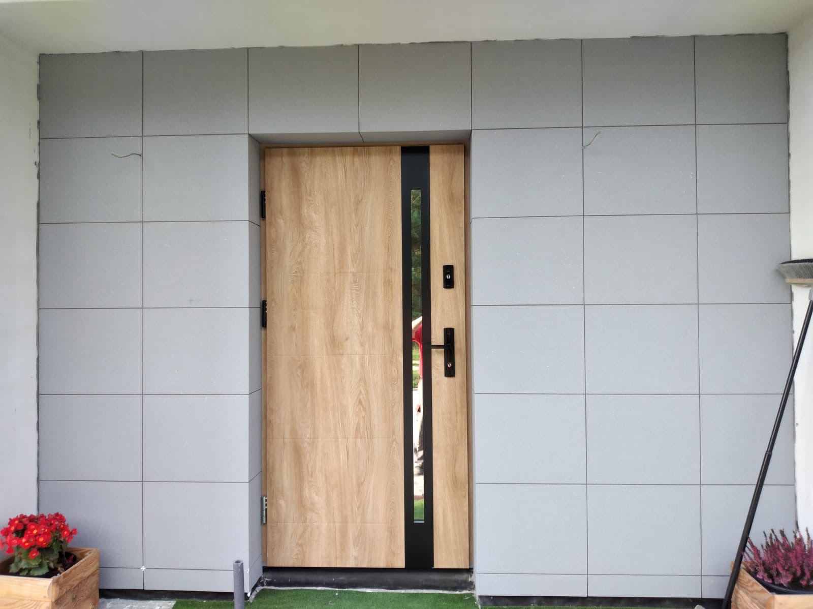 Elewacja domu wykończona szarymi płytami imitującymi kamień, z drewnianymi drzwiami wejściowymi z czarnymi akcentami. Widoczne kwiaty w donicach i fragment trawnika.