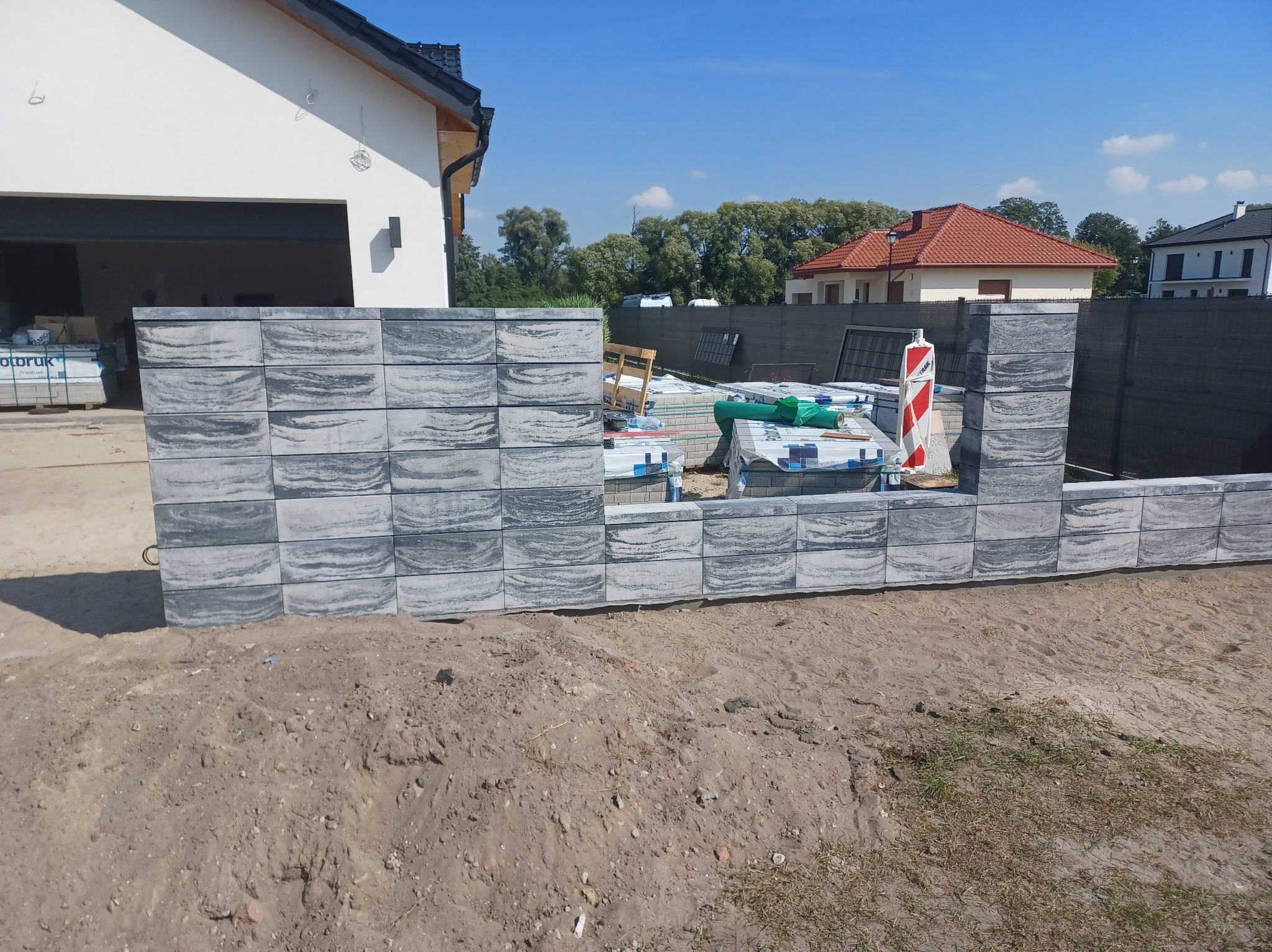 Wznoszony mur z szarych, wzorzystych bloczków betonowych na tle budynków i materiałów budowlanych. Widoczny fragment ogrodzenia panelowego.