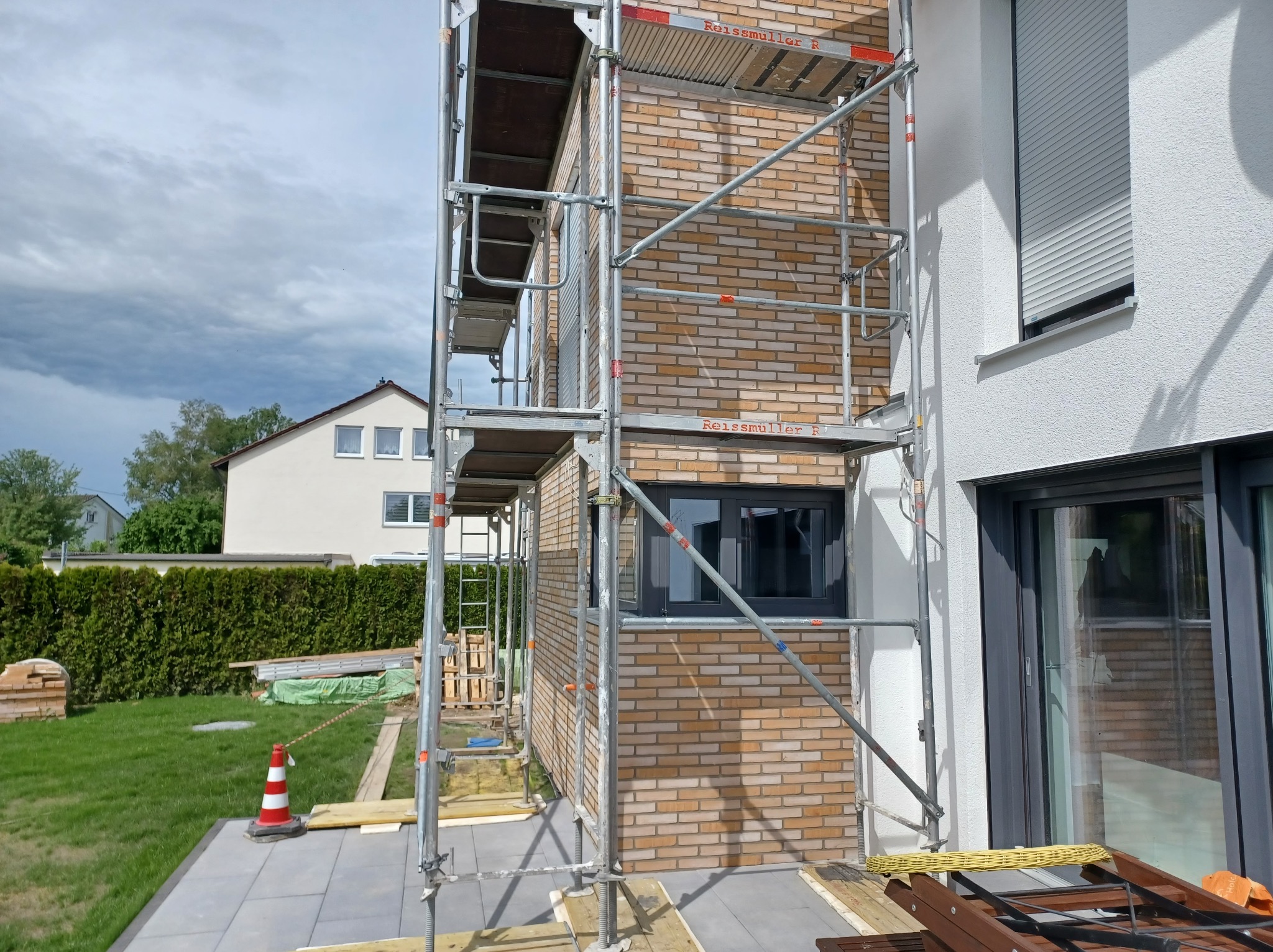 Ściana budynku w trakcie murowania z cegły, widoczne rusztowanie, okno z roletą i fragment ogrodu. Prace murarskie w toku, ceglana elewacja w odcieniach beżu i brązu.