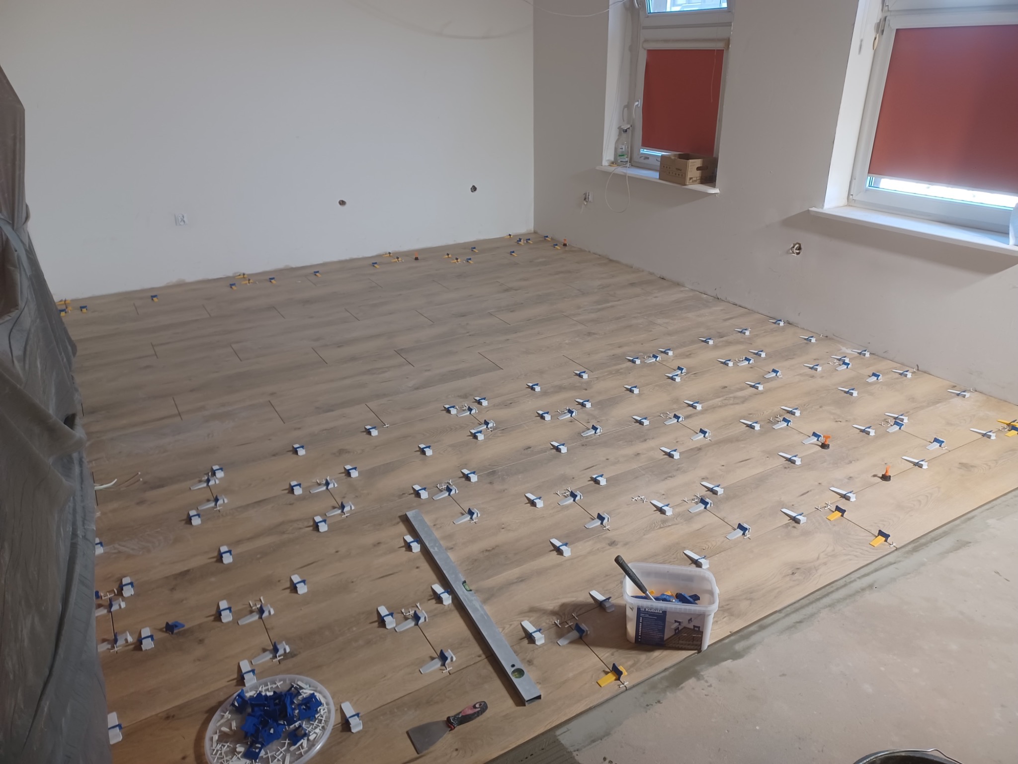 Układanie paneli podłogowych w jasnym odcieniu drewna, z użyciem klipsów poziomujących. Widoczna poziomica i pojemnik z klipsami. Dwa okna z roletami w tle.