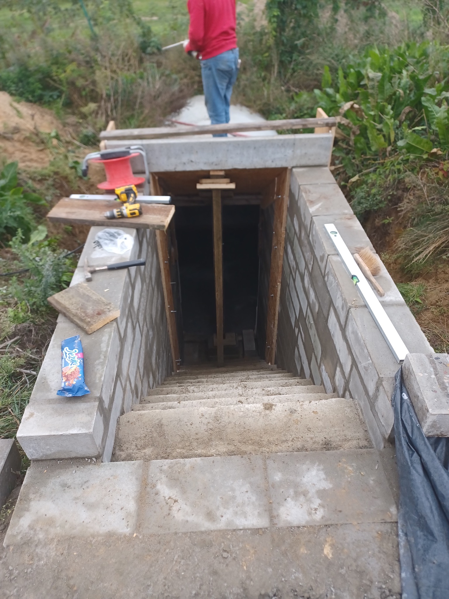 Betonowe schody prowadzące w dół, z widocznymi narzędziami murarskimi i poziomnicą. W tle osoba w czerwonej bluzie i zielona roślinność.