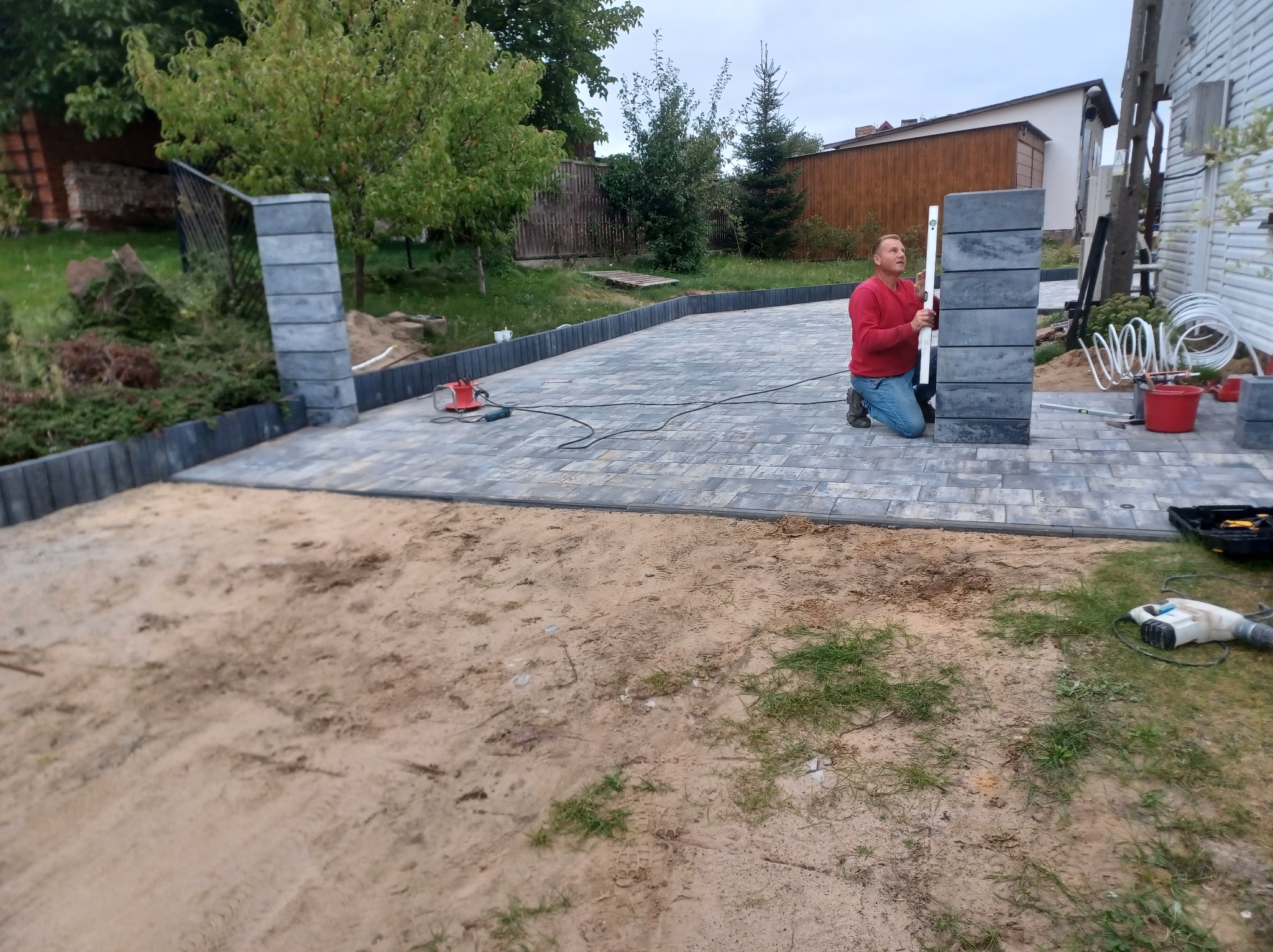 Układanie kostki brukowej i montaż obrzeży w Stolno. Mężczyzna z poziomicą kontroluje pionowość słupka ogrodzeniowego z szarej kostki. Widoczne narzędzia i częściowo ułożony chodnik.