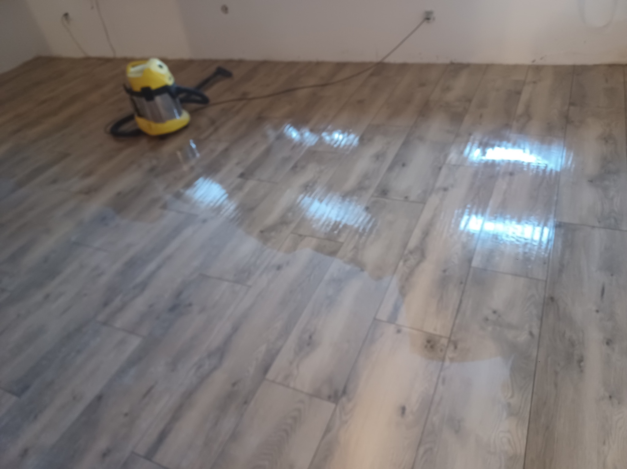 Położona podłoga z paneli imitujących drewno, mokra po umyciu. W rogu żółty odkurzacz przemysłowy. Widoczne gniazdka elektryczne na ścianie.