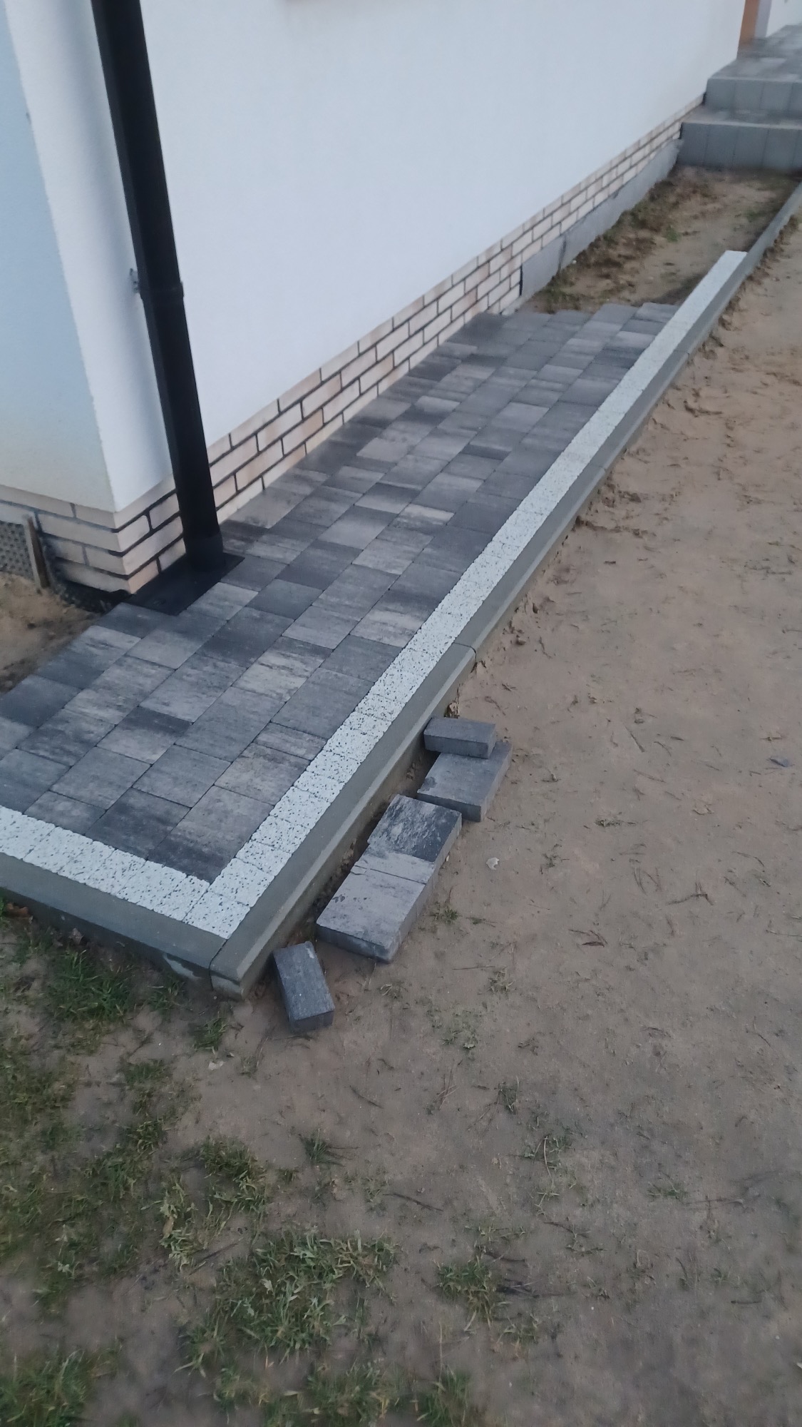 Ułożona ścieżka z kostki brukowej w odcieniach szarości, obrzeża z jasnej kostki. Część kostki leży obok ścieżki. Widoczny fragment elewacji budynku.