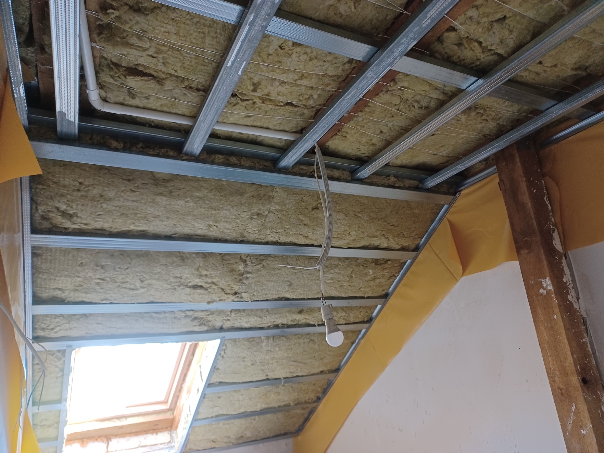 Skośny sufit poddasza z izolacją z wełny mineralnej, widoczne metalowe profile i okno dachowe. Żółta folia paroizolacyjna na ścianie. W tle drewniany element konstrukcyjny.