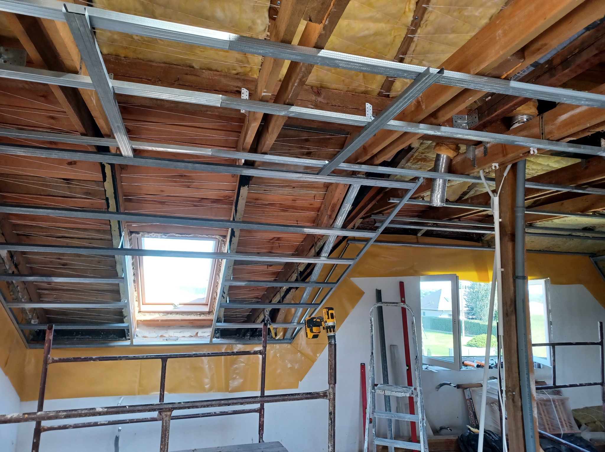 Szkielet sufitu podwieszanego z profili metalowych na poddaszu, widoczne ocieplenie wełną mineralną, okno dachowe i drabina. Prace wykończeniowe w toku.