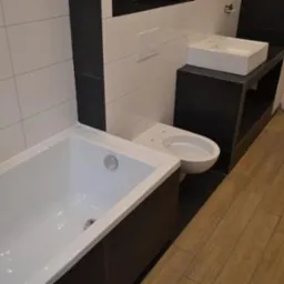 Nowoczesna aranżacja łazienki z białą wanną, toaletą i umywalką na czarnym blacie. Jasne płytki na ścianach i drewnopodobna podłoga tworzą spójną całość.