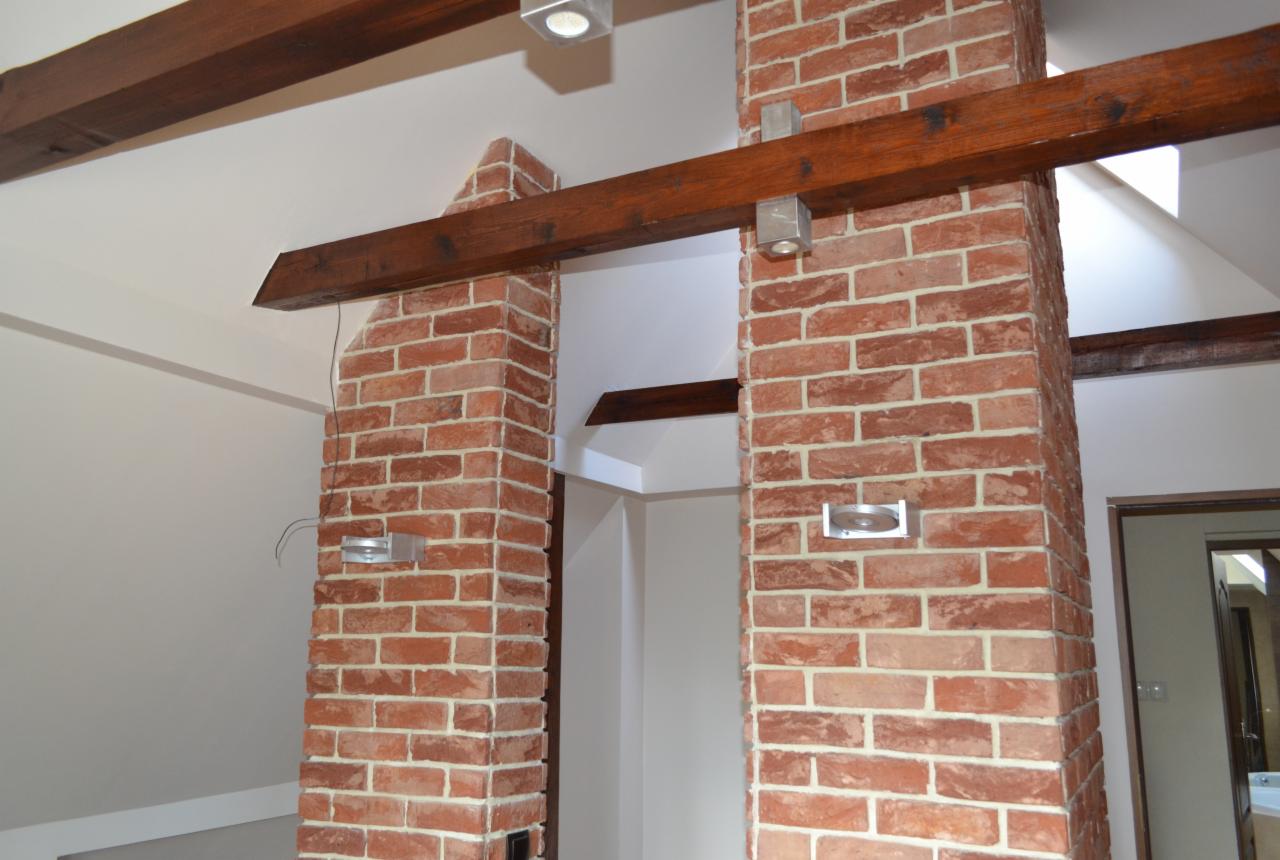 Wykończone cegłą filary z drewnianymi belkami stropowymi i nowoczesnym oświetleniem, tworzące unikalny element dekoracyjny wnętrza.