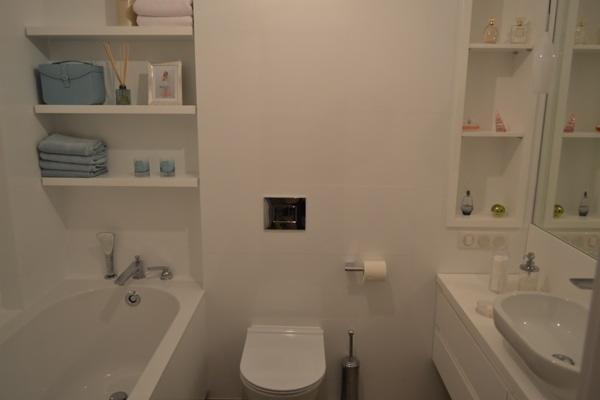 Jasna łazienka z wanną, toaletą i umywalką. Na ścianie wnęki z półkami i dekoracjami. Minimalistyczny design, białe wykończenie, nowoczesna armatura.