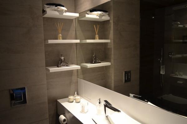 Nowoczesna łazienka z szarymi płytkami, białymi półkami z dekoracjami i minimalistyczną umywalką. Lustro powiększa przestrzeń, a oświetlenie podkreśla detale.