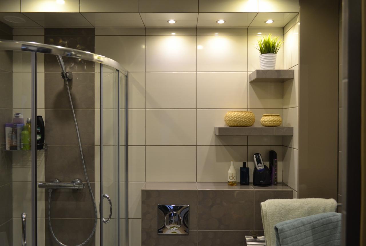 Nowoczesna łazienka z kabiną prysznicową, beżowymi płytkami i półkami z dekoracjami. Oświetlenie sufitowe podkreśla aranżację.