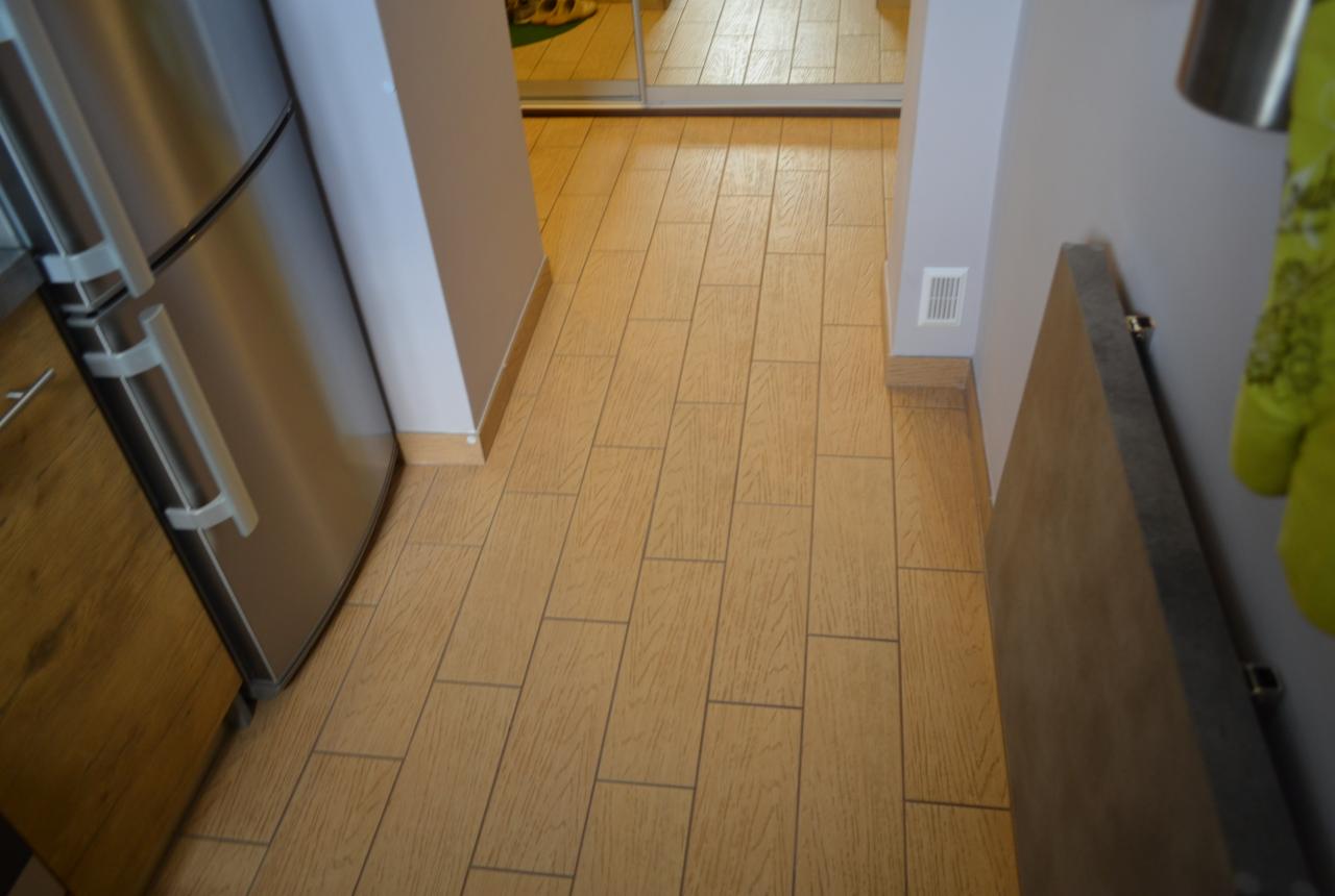 Ułożone płytki podłogowe imitujące drewno w korytarzu, widok z perspektywy wejścia, z fragmentem lodówki po lewej i grzejnikiem po prawej.