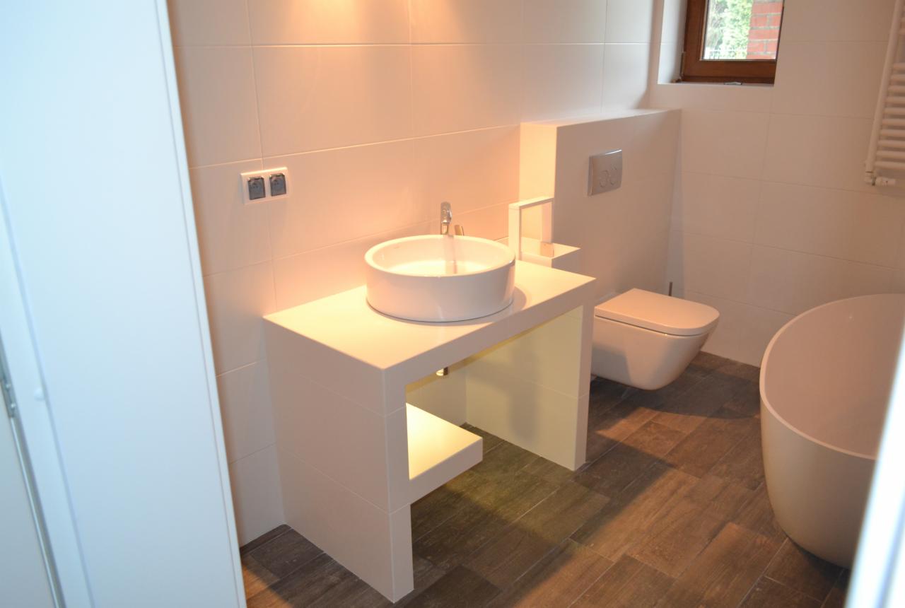 Nowoczesna łazienka z białą, okrągłą umywalką na blacie z półką, wiszącą toaletą i wolnostojącą wanną. Płytki na ścianach i podłodze w odcieniach bieli i brązu.