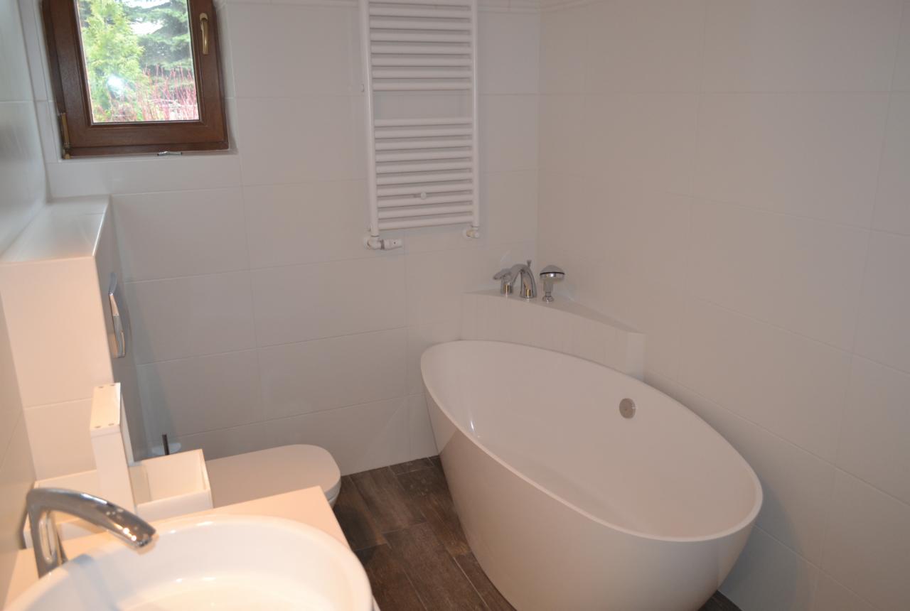 Biała, owalna wanna wolnostojąca w nowoczesnej łazience z białymi płytkami, drewnopodobną podłogą i grzejnikiem drabinkowym. Okno z drewnianą ramą.