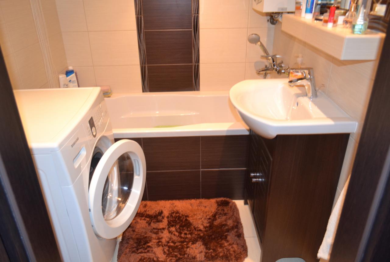 Nowoczesna łazienka z pralką, umywalką na szafce i wanną w brązowo-beżowej kolorystyce. Widoczny dywanik i elementy armatury. Neutralne oświetlenie.