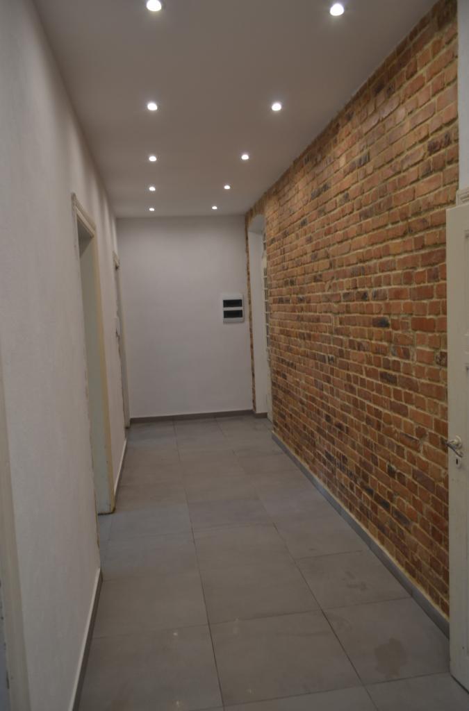 Korytarz po remoncie: ściana z cegły, szare płytki podłogowe, białe ściany i sufit z oświetleniem punktowym. Widok z perspektywy.