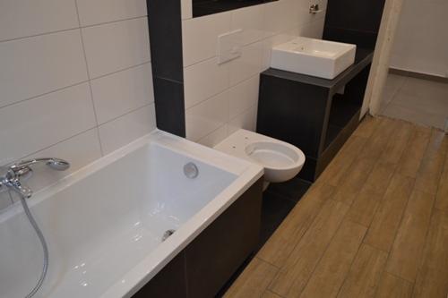 Nowoczesna aranżacja łazienki z białą wanną, toaletą i umywalką na czarnym blacie. Jasne płytki na ścianach i drewnopodobna podłoga tworzą spójną całość.