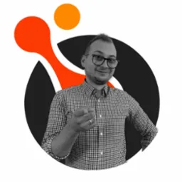 Czarno-białe zdjęcie mężczyzny w okularach i koszuli w kratę, wskazującego palcem w kierunku widza, na tle abstrakcyjnego logo w kolorach pomarańczowym, białym i czarnym.