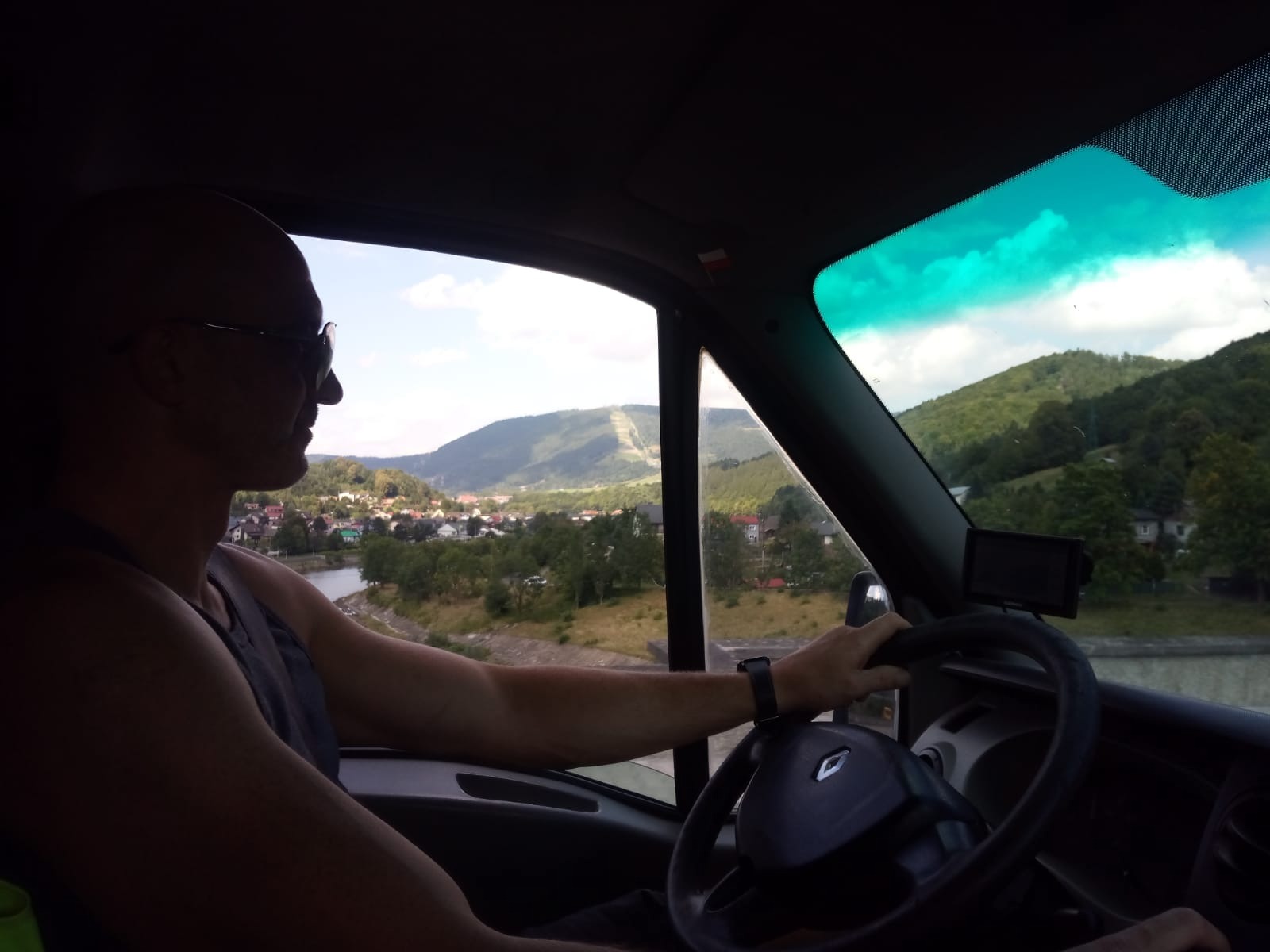 Kierowca w okularach prowadzi Renault Master z widokiem na górski krajobraz i rzekę, wnętrze kabiny z zieloną szybą.