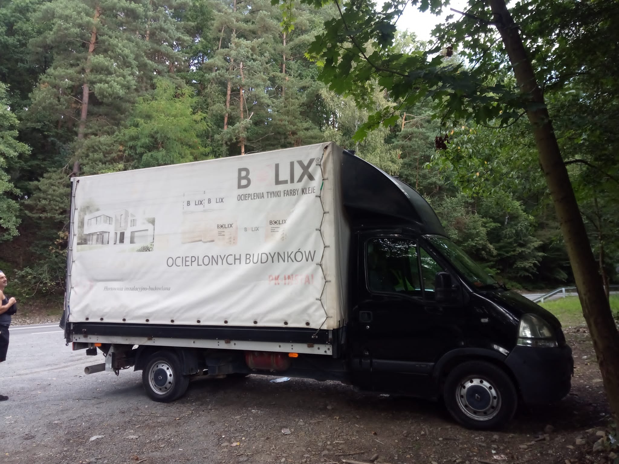 Czarny bus z plandeką Bolix zaparkowany na poboczu, w tle las. Na plandece reklama ociepleń, tynków, farb i klejów.