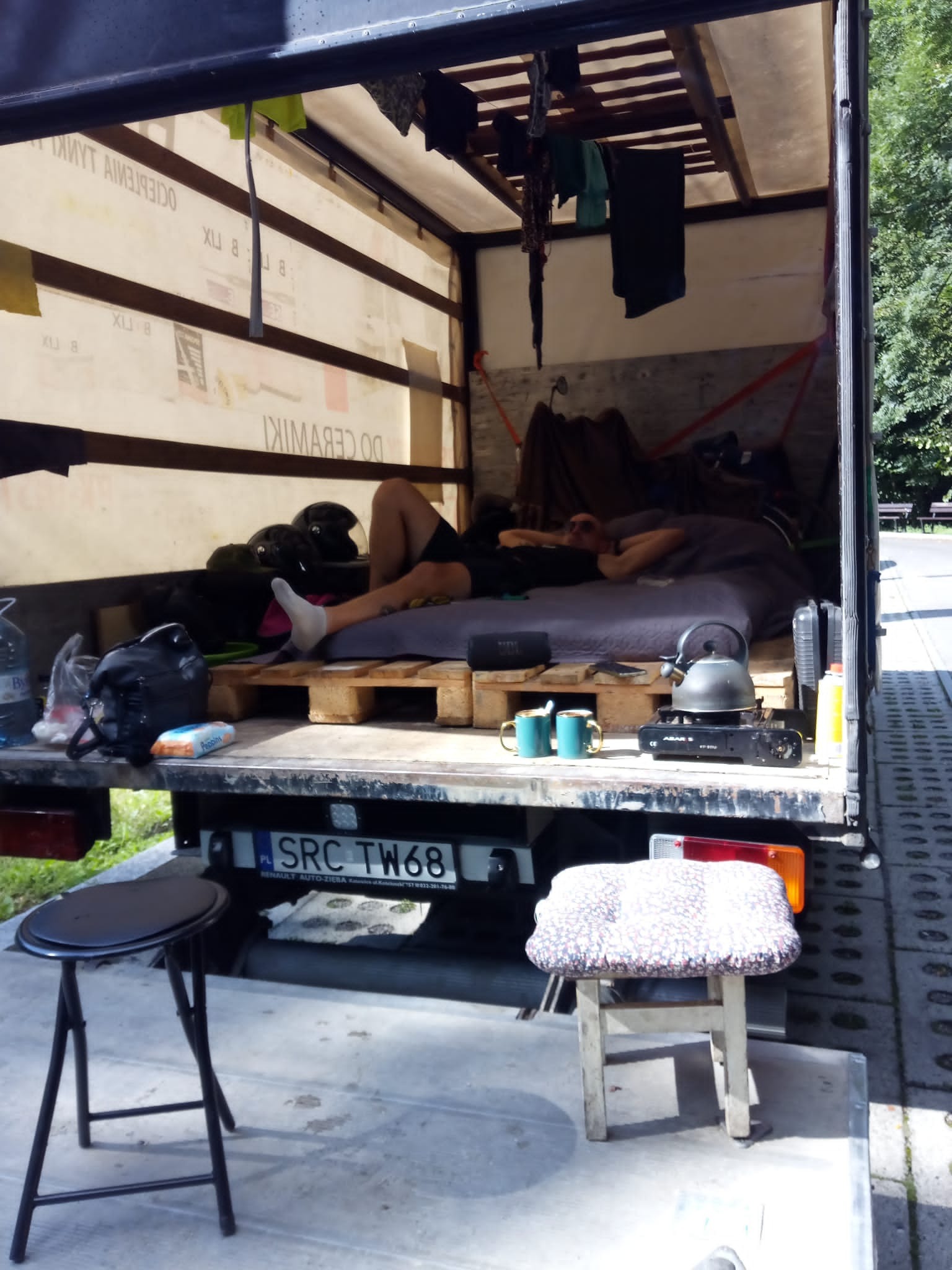 Wnętrze furgonetki zaadaptowane na tymczasowe mieszkanie podczas transportu: łóżko na paletach, kuchenka turystyczna, naczynia, ubrania suszące się pod sufitem, tablice rejestracyjne SRC TW68.