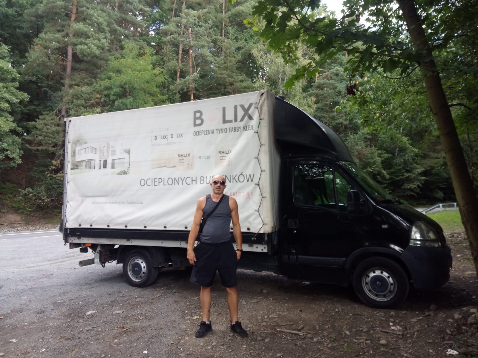 Mężczyzna w okularach przeciwsłonecznych stoi przed czarnym busem z reklamą materiałów budowlanych Bolix na plandece, w tle las.