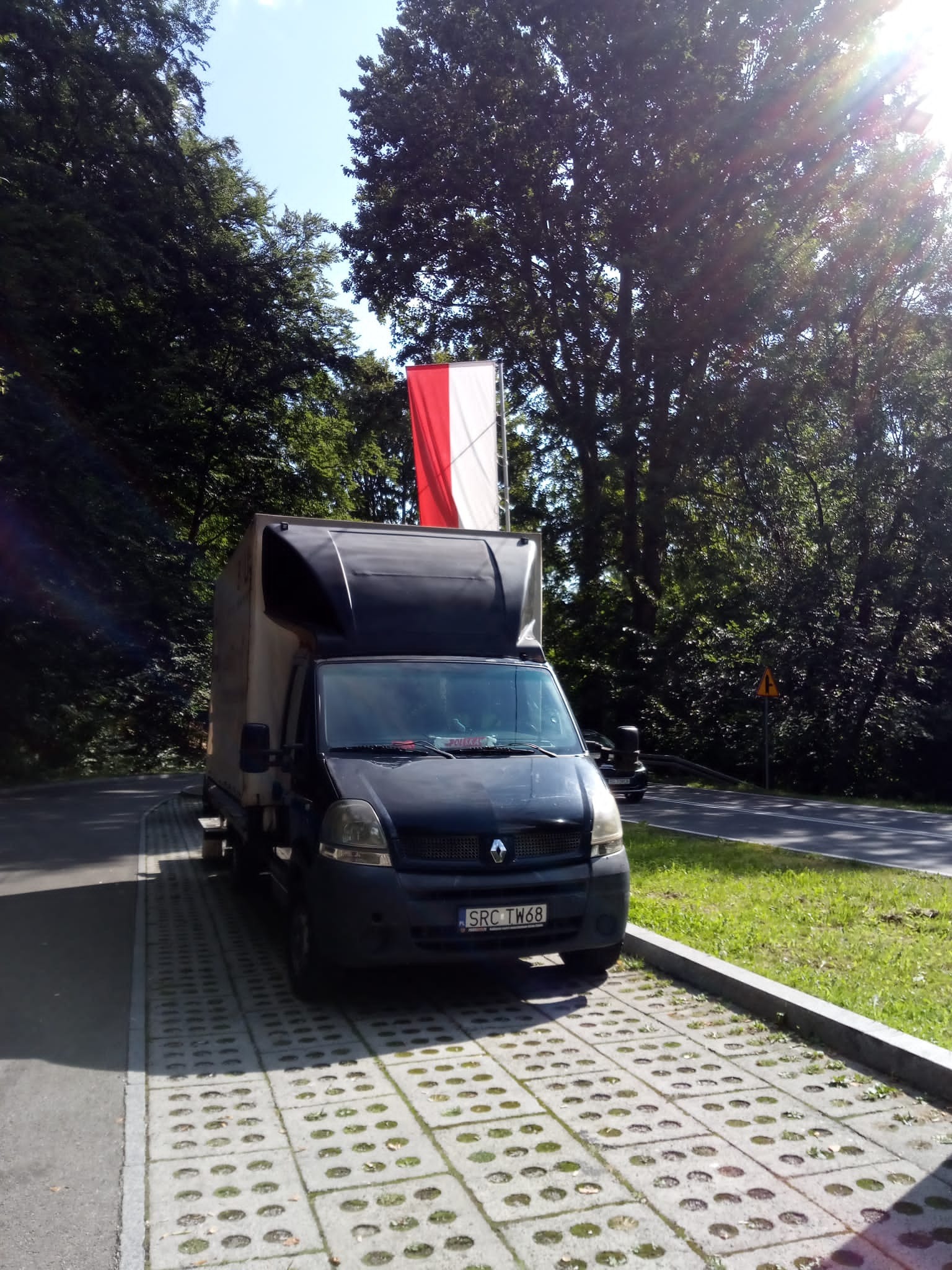 Czarny bus dostawczy Renault Master z polską flagą na dachu, zaparkowany na betonowej kostce w otoczeniu drzew, widoczna tablica rejestracyjna SRC TW68.