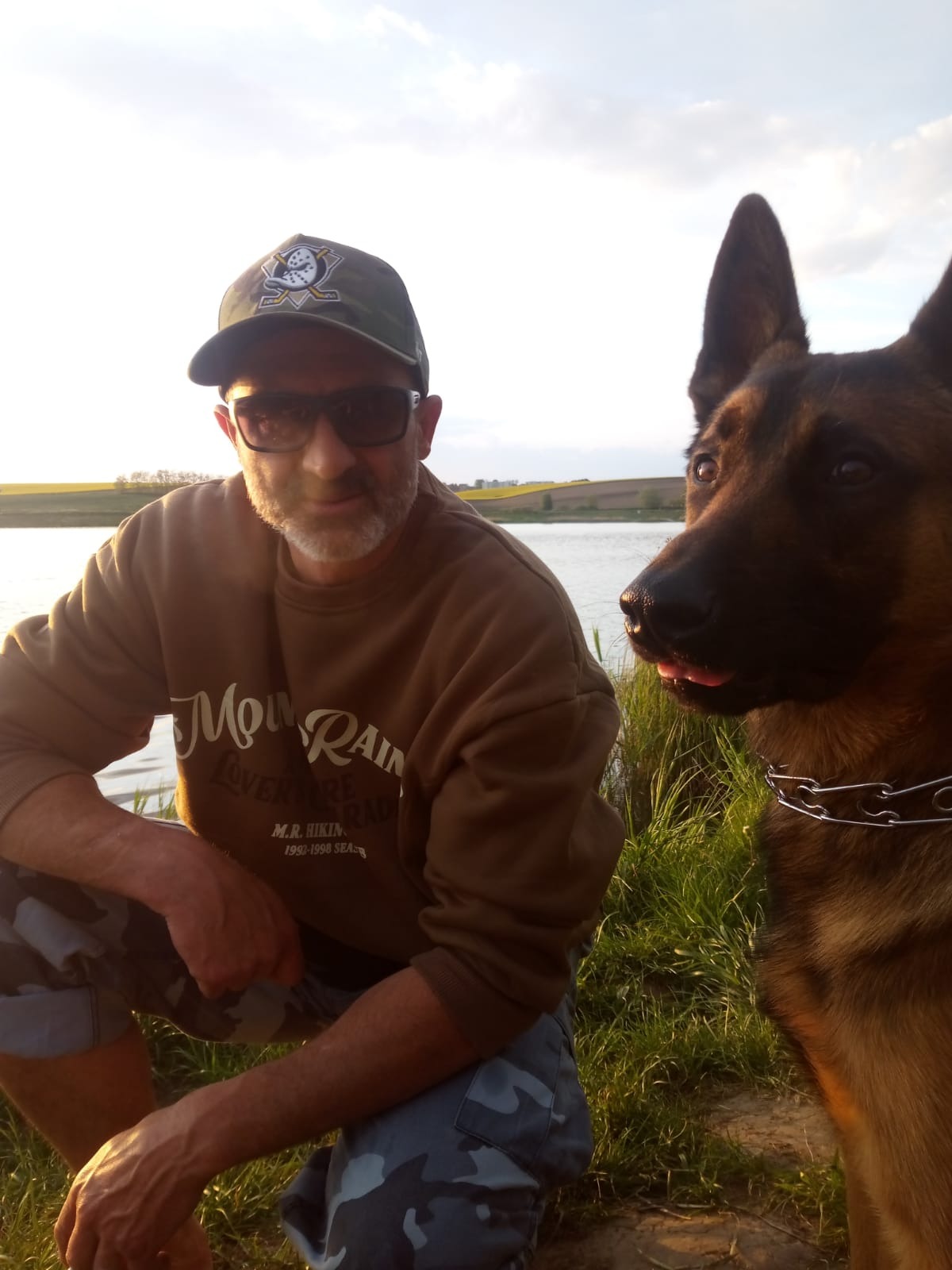 Mężczyzna w okularach i czapce kuca obok owczarka belgijskiego na tle jeziora i łąki w słoneczny dzień. Pies ma otwarty pysk.