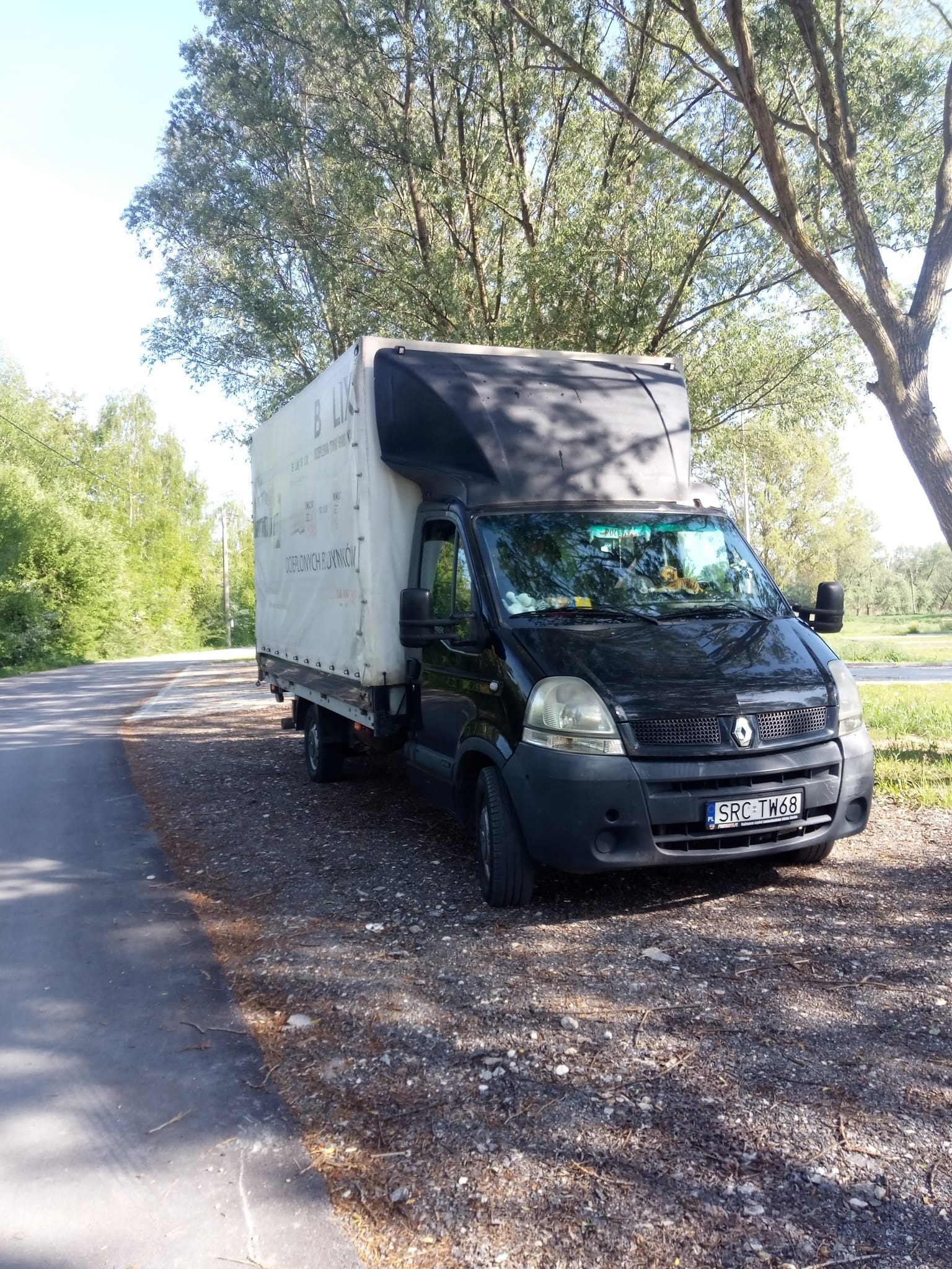 Czarny bus Renault Master z plandeką, zaparkowany na poboczu drogi w otoczeniu drzew. Na plandece widoczny napis reklamowy, a na desce rozdzielczej pluszowe zabawki.