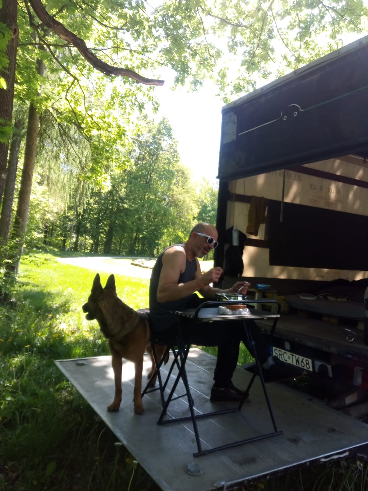 Przerwa w transporcie: mężczyzna w okularach je posiłek na platformie busa, obok pies owczarek, w tle zielony park i tablica rejestracyjna pojazdu.