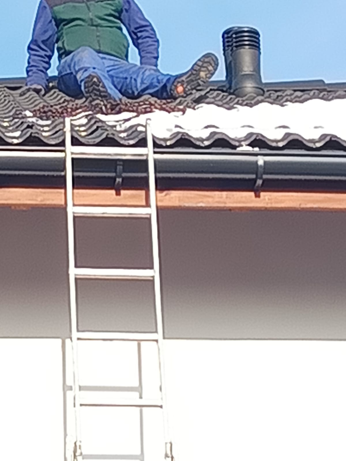Dekarz siedzi na dachu obok komina, opierając nogi o drabinę. Widoczne dachówki, rynna i fragment ściany budynku. Ujęcie z dołu.