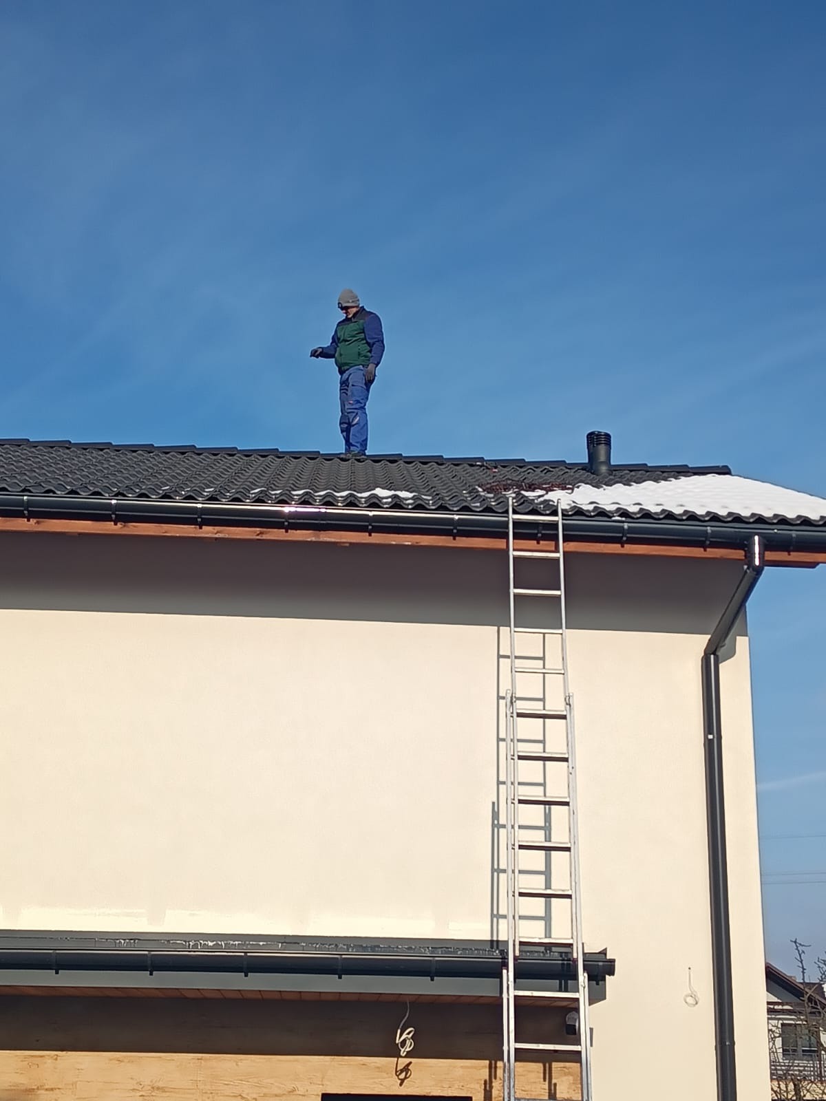 Pracownik w kombinezonie na dachu domu z czarną dachówką, obok komin i drabina. Częściowo ośnieżony dach w słoneczny dzień. Praca na wysokościach.