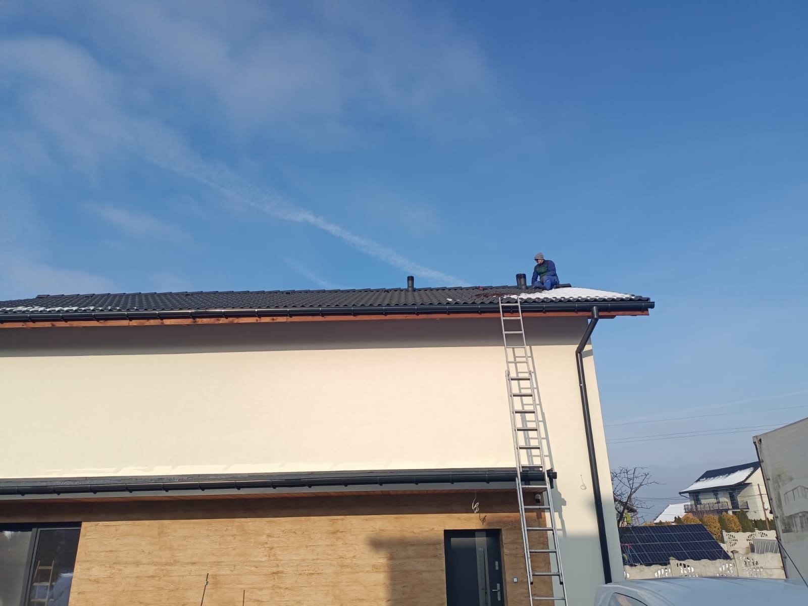 Praca na dachu: dekarz w niebieskim kombinezonie siedzi na czarnym dachu domu z kominem, obok drabina. Częściowo ośnieżony dach i błękitne niebo w tle.